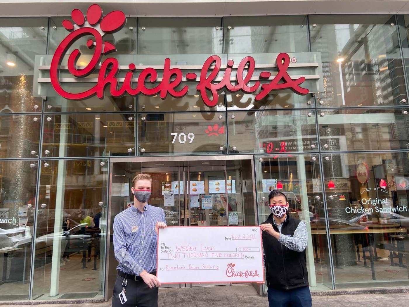Two men holding a large check in front of a Chick-fil-A restaurant entrance.