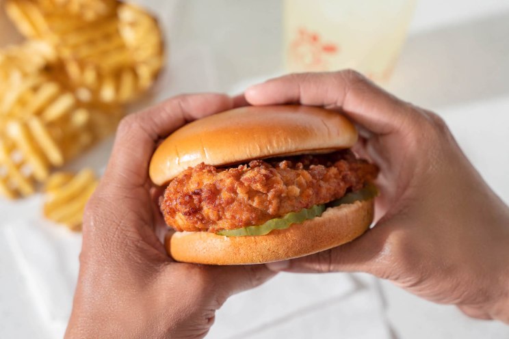 Close-up of a Chick-fil-A Chicken Sandwich, held by two hands, with waffle fries and a drink in the background.
