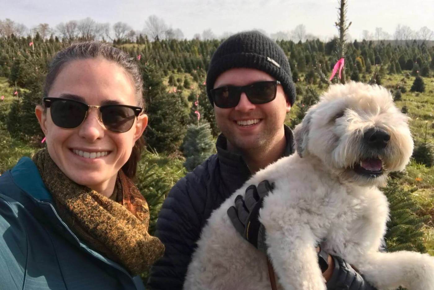 A man and woman in sunglasses pose outdoors at a Christmas tree farm, the man holding a fluffy white dog.
