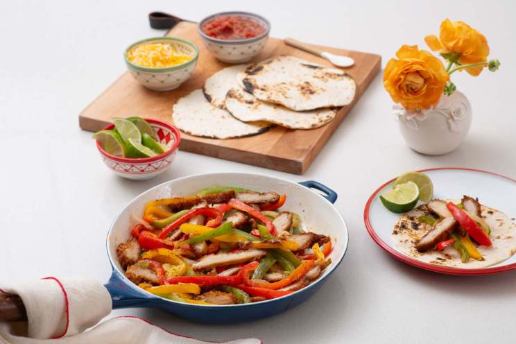 Fajitas with sliced chicken, colorful bell peppers, and accompaniments including tortillas, salsa, cheese, lime, and yellow flowers.