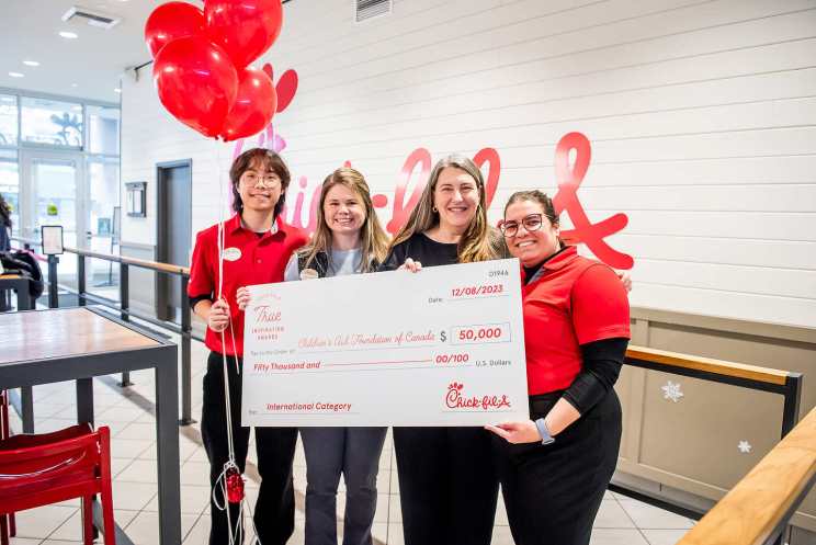 Four people stand together holding a large check and red balloons in a bright, modern interior.