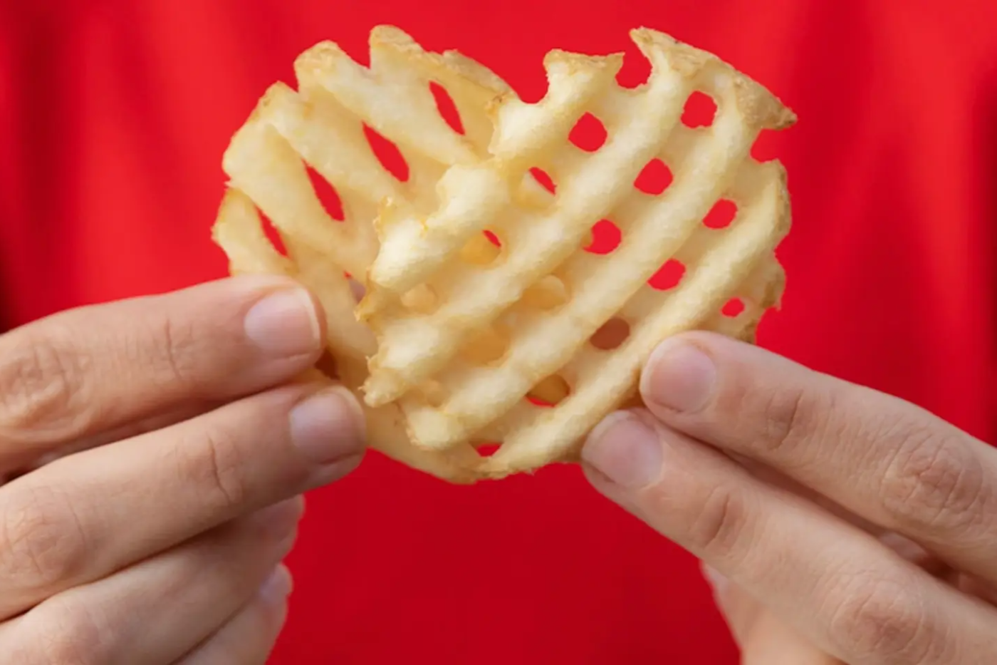 A waffle fry resembling a heart, held between two hands with a red background.