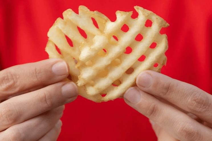 A waffle fry resembling a heart, held between two hands with a red background.