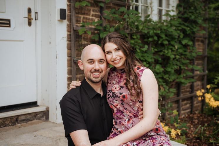 A couple smiling together on a stone step in front of a house, surrounded by ivy and flowers.