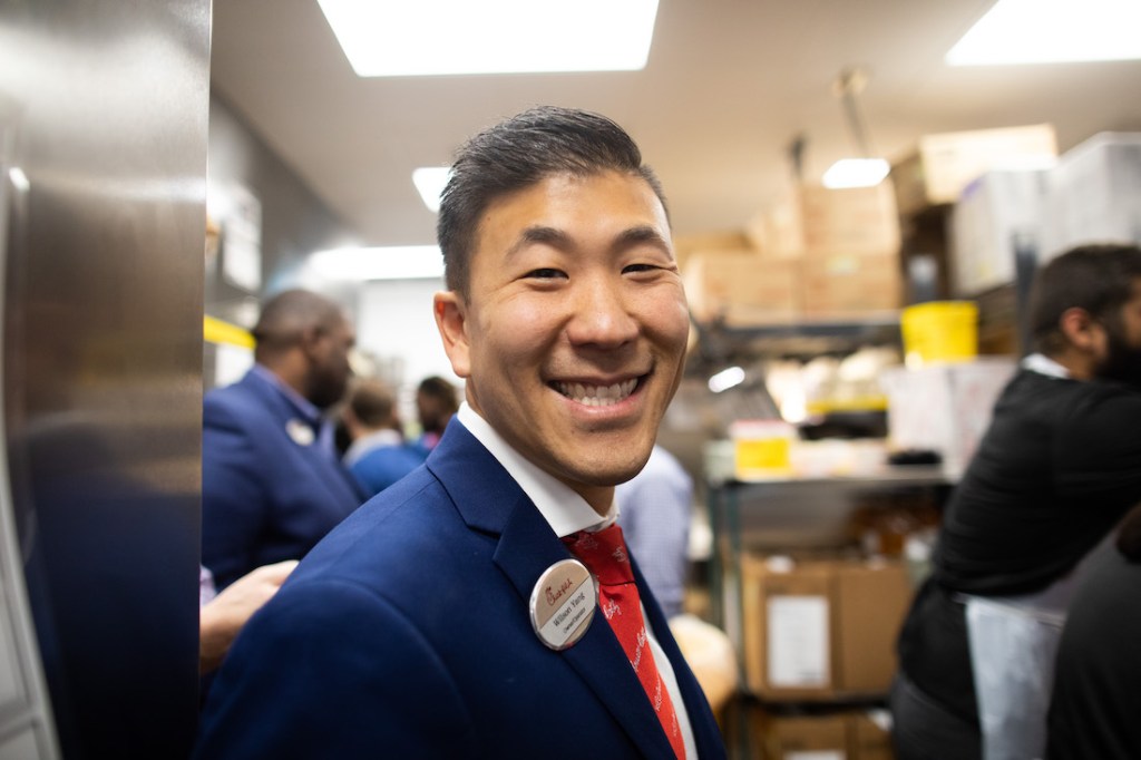 Operator Wilson Yang smiling in a Chick-fil-A kitchen, wearing a suit and name badge, surrounded by team members.