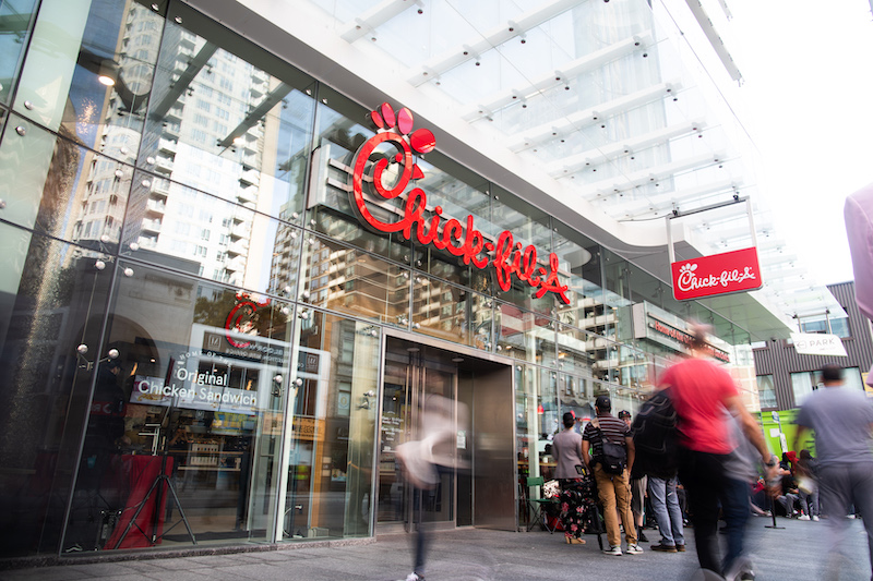 Customers lining up outside a glass-front Chick-fil-A restaurant in Canada, with city buildings reflected in the windows.