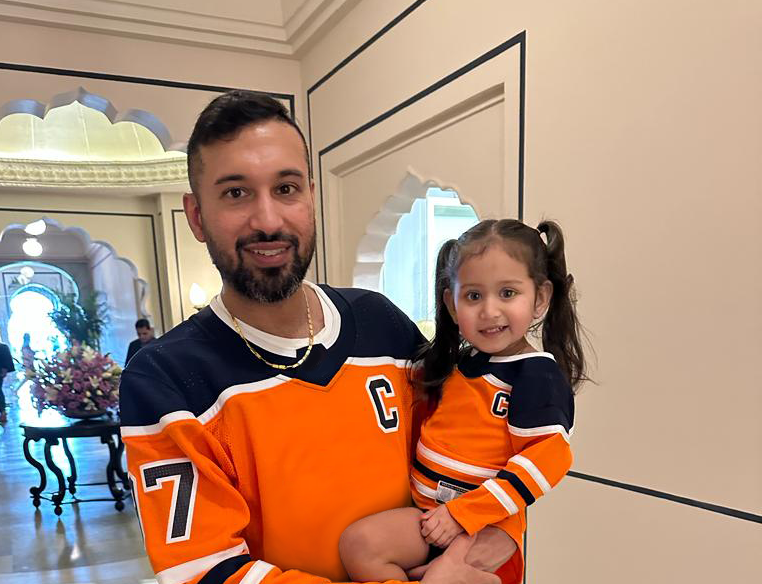 Jag Dhanju holding a child, both in matching orange sports jerseys, standing indoors with ornate architecture in the background.