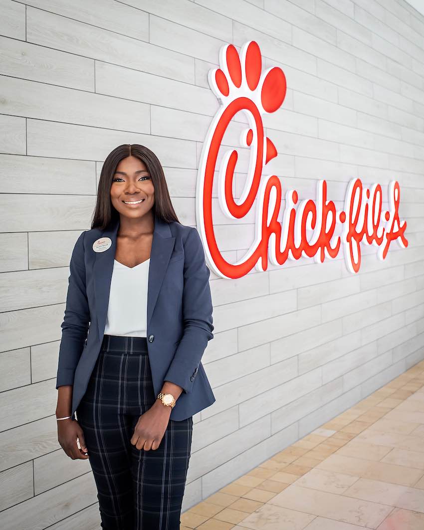 Mufuti Sanusi in business attire standing in front of a "Chick-fil-A" logo on a wooden paneled wall.