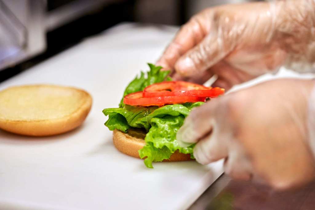 A person adding lettuce, tomatoes and pickles to a bun.