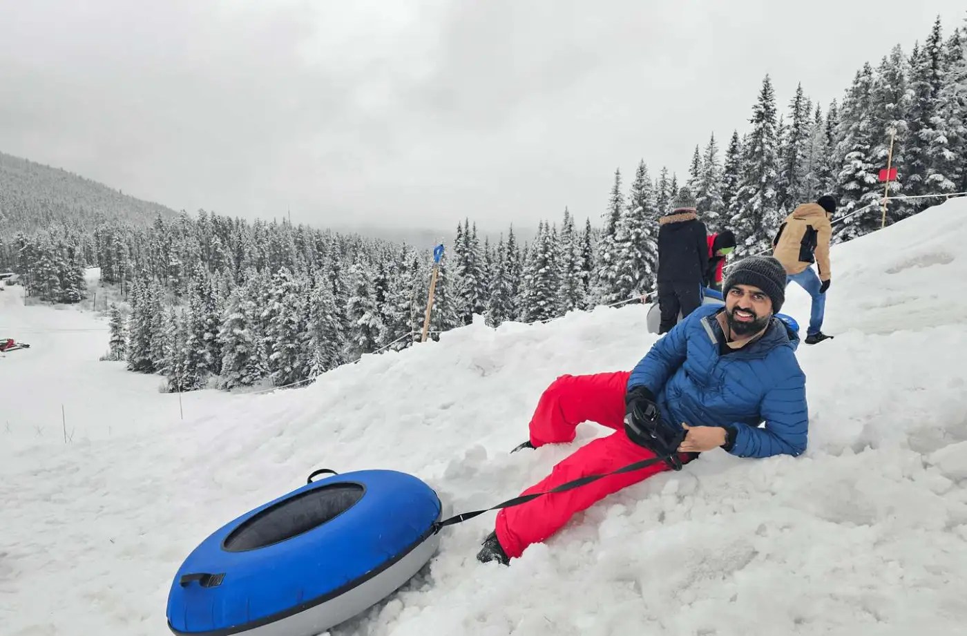 Person lying on snowy ground with a snow tube and forest in the background.