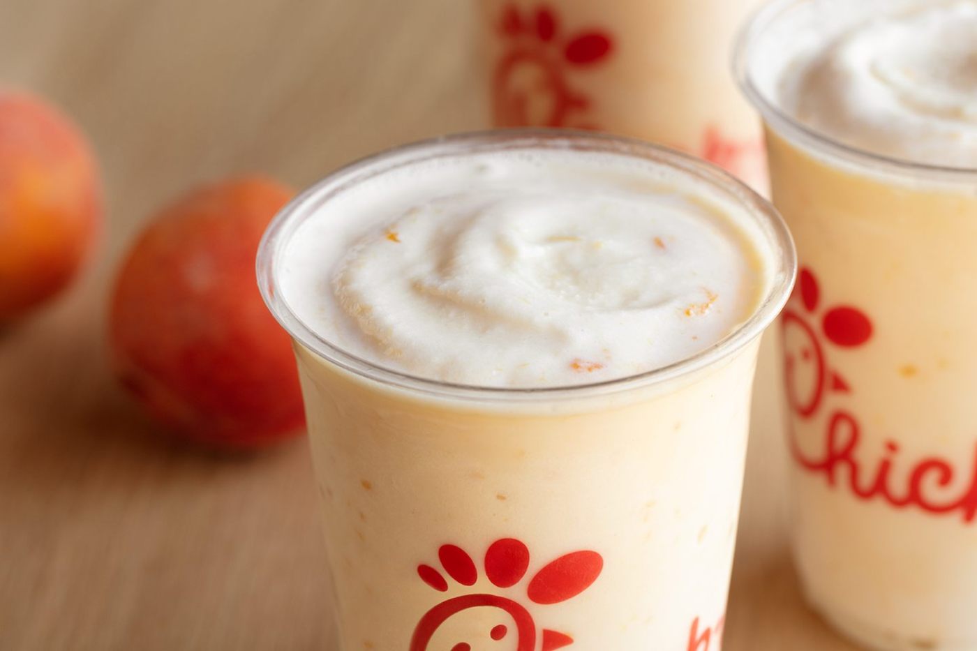 Three cups with peach shakes, accompanied by two peaches on a wooden surface.