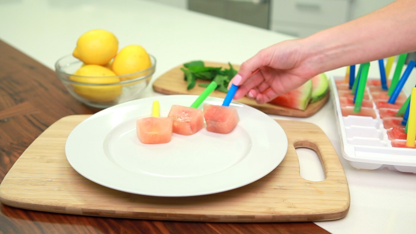 A hand places a pink popsicle with a blue stick on a white plate next to other popsicles, with lemons and mint in the background.