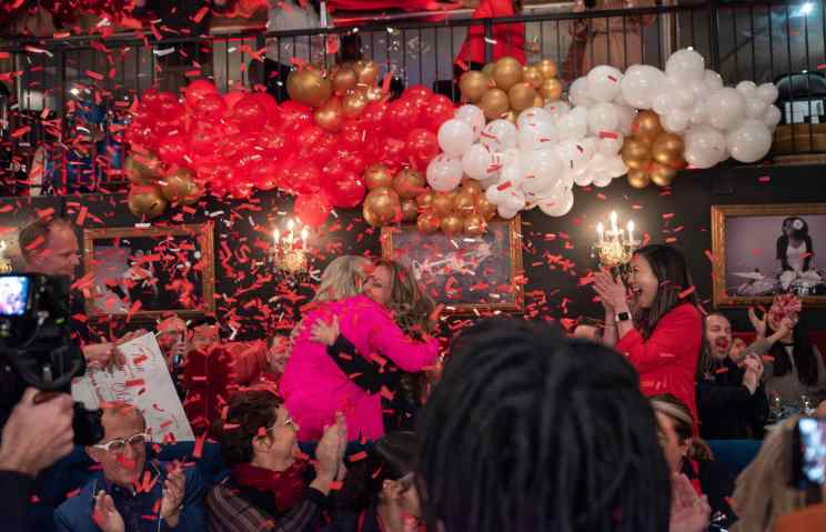 True Inspiration Award celebration with red, white, and gold balloons and confetti. Two women embrace while others clap around them.