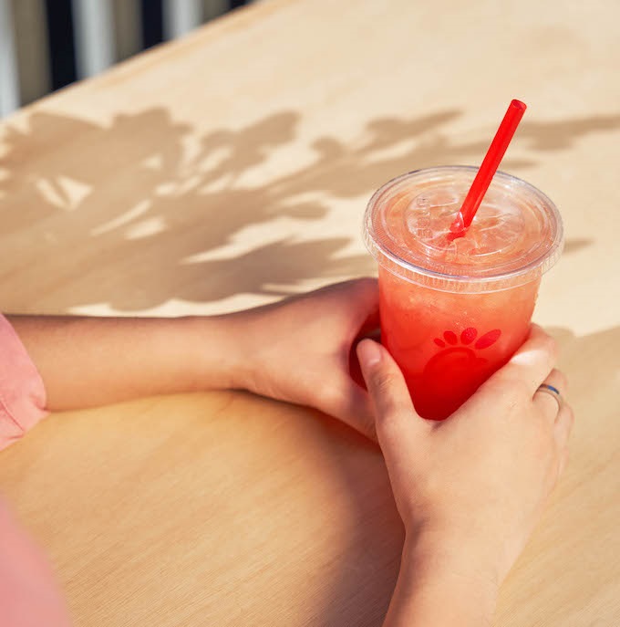 Person holding a clear cup with Sunjoy and red straw on a wooden table with leaf shadows.