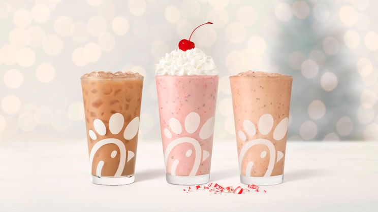 Glasses of Peppermint Chip Milkshake, Peppermint Iced Coffee, and Peppermint Chip lined up in a row, sitting next to pieces of peppermint, with twinkling holiday lights in the background