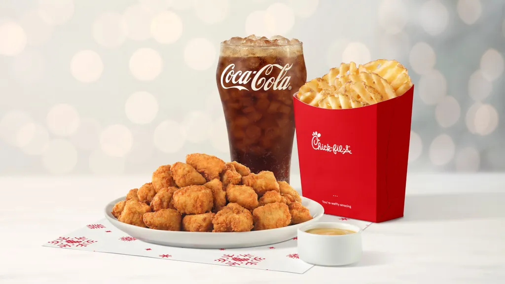30-ct Chick-fil-A Nuggets sitting on holiday-themed parchment paper, next to a glass of Coca-Cola® and a carton of Waffle Fries with twinkling holiday lights in the background