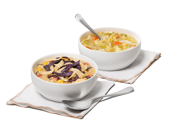 A bowl of Chicken Tortilla Soup on a cloth napkin next to a bowl of Chicken Noodle Soup on a cloth napkin