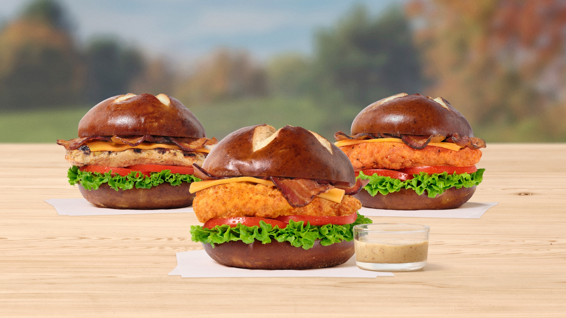 Three Pretzel Cheddar Club Sandwiches, one with the Original filet, one with the Grilled filet and one with Spicy filet all on white napkins next to a small glass bowl of Creamy Dijon Mustard Sauce with a window in the
background