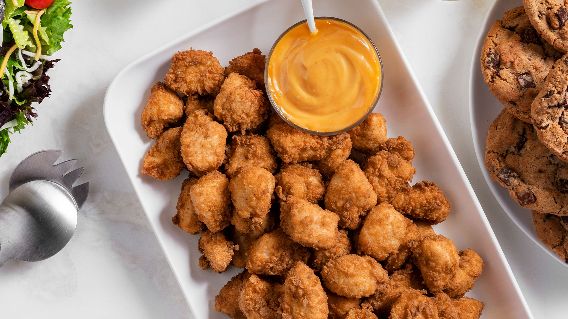 Chick-fil-A Nugget Tray on a white platter with a small bowl of Chick-fil-A Sauce and Chocolate Chunk Cookie Tray on a white plate, and a Garden Salad Tray